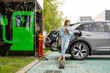 Young woman stands with a smart phone, waiting for her electric car to charge on a public charging station outdoors. Concept of modern lifestyle and green energy for transportation