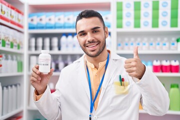 Canvas Print - Young hispanic man working at pharmacy drugstore holding pills smiling happy and positive, thumb up doing excellent and approval sign