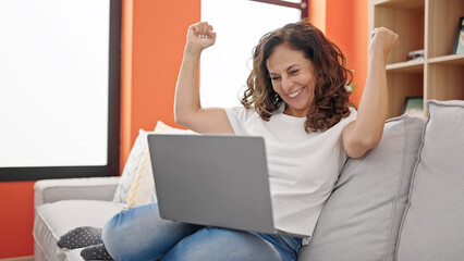 Canvas Print - Middle age hispanic woman using laptop sitting on sofa with winner expression at home