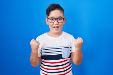 Poster - Young hispanic kid standing over blue background celebrating surprised and amazed for success with arms raised and open eyes. winner concept.