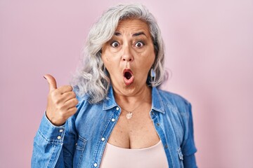 Poster - Middle age woman with grey hair standing over pink background surprised pointing with hand finger to the side, open mouth amazed expression.