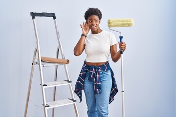 Poster - African american woman holding roller painter smiling with hand over ear listening an hearing to rumor or gossip. deafness concept.