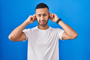 Poster - Young hispanic man standing over blue background covering ears with fingers with annoyed expression for the noise of loud music. deaf concept.