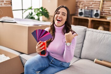 Sticker - Young brunette woman moving to a new home choosing wall painting pointing thumb up to the side smiling happy with open mouth