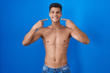 Poster - Young hispanic man standing shirtless over blue background smiling cheerful showing and pointing with fingers teeth and mouth. dental health concept.