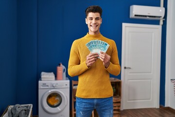 Sticker - Young hispanic man at laundry room holding brazilian reals smiling with a happy and cool smile on face. showing teeth.