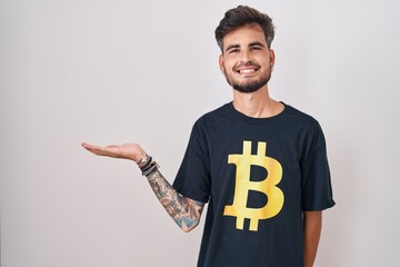 Sticker - Young hispanic man with tattoos wearing bitcoin t shirt smiling cheerful presenting and pointing with palm of hand looking at the camera.