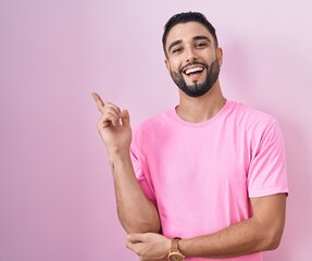 Wall Mural - Hispanic young man standing over pink background with a big smile on face, pointing with hand finger to the side looking at the camera.