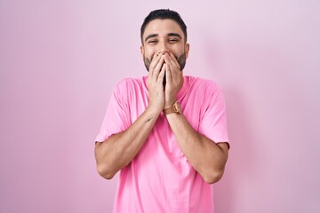 Poster - Hispanic young man standing over pink background laughing and embarrassed giggle covering mouth with hands, gossip and scandal concept