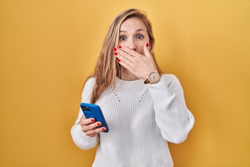 Poster - Young blonde woman using smartphone typing message shocked covering mouth with hands for mistake. secret concept.