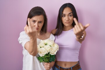 Wall Mural - Hispanic mother and daughter holding bouquet of white flowers showing middle finger, impolite and rude fuck off expression