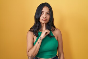 Sticker - Brunette woman standing over yellow background thinking concentrated about doubt with finger on chin and looking up wondering