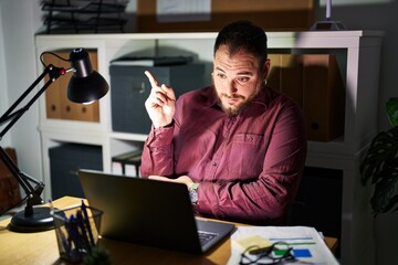 Wall Mural - Plus size hispanic man with beard working at the office at night with a big smile on face, pointing with hand and finger to the side looking at the camera.