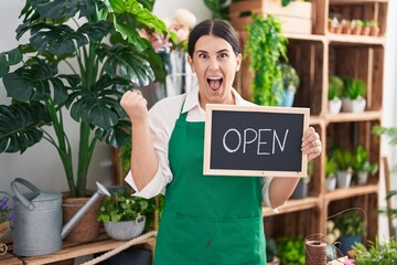 Wall Mural - Young hispanic woman working at florist holding open sign screaming proud, celebrating victory and success very excited with raised arm