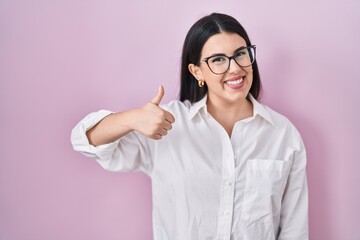 Sticker - Young brunette woman standing over pink background doing happy thumbs up gesture with hand. approving expression looking at the camera showing success.