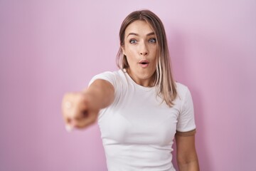 Poster - Blonde caucasian woman standing over pink background pointing with finger surprised ahead, open mouth amazed expression, something on the front