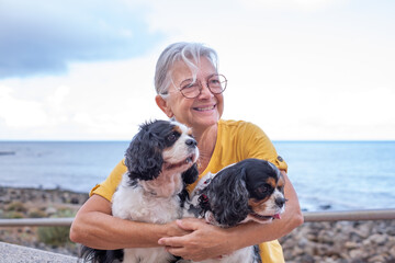 smiling senior woman in yellow jersey sitting close to the beach embracing her two cavalier king cha