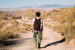 Young man walk away from camera, in leather boots, wear black backpack and carry water flask on empty dusty desert road. Moody solo adventure. Hot sunny day for a hike