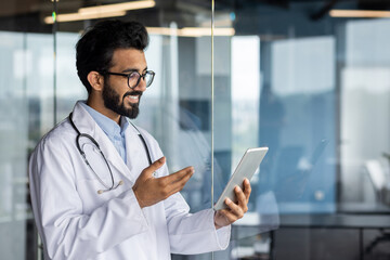 Successful professional Indian doctor working inside modern clinic, man consulting patients remotely, using tablet computer for video call, and long distance communication