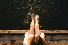 Woman Relaxes By The Lake Sitting On The Edge Of A Wooden Jetty, Swing Feet Near The Water Surface, Top View.