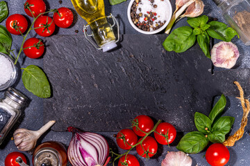 Black cooking background with useful cooking italian Mediterranean ingredients - tomatoes, basil leaves, greens, olive oil, salt, pepper, garlic, flat lay black concrete table top view copy space