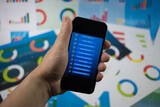 Fototapeta  - Ręka trzymająca telefon z niebieskim ekranem na tle wykresów.