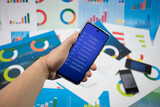 Fototapeta  - Ręka trzymająca telefon z niebieskim ekranem z napisami na tle wykresów.