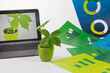 Mała roślina w doniczce obok komputera z rośliną na ekranie wśród zielonych i niebieskich plansz z wykresami.