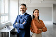 Successful businessman and businesswoman standing back to back with arms crossed and smiling at camera, office interior