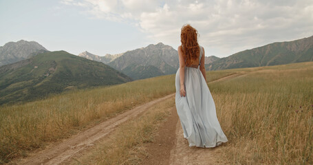 Wall Mural - Girl with red hair walking down the wheel trails on meadow with scenic mountain background - freedom, tranquility concept .Copy space