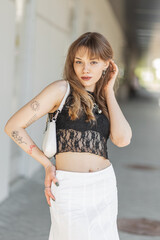 Wall Mural - Beautiful young stylish hipster woman with a tattoo on her arms in a fashionable summer streetwear with a lace top, a skirt and a handbag walks in the city and looks at the camera