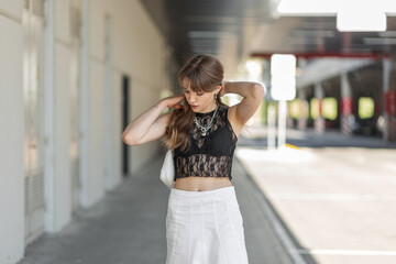 Wall Mural - Stylish beautiful young woman in a fashionable black lace top with a bra and a white skirt with a handbag walks in the city and straightens her hair