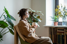 Thoughtful Dreaming Creative Girl Sits On Armchair With Cup Coffee Look Out Window Enjoy Day Time In Cozy Place House With Green Plants In Pots. Young Woman Thought About Life. Mental Health Concept.