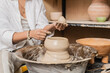 © LIGHTFIELD STUDIOS - Cropped view of young female artisan in workwear holding and molding clay on pottery wheel and working near bowl in ceramic workshop, clay sculpting process concept
