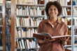 © AnnaStills - Portrait of African American student smiling at camera while reading book in library