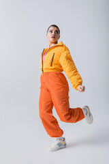 stylish look, outerwear, casual attire, fashion model posing in yellow puffer jacket and orange pants on grey background, woman with short hair running and looking away, modern subculture