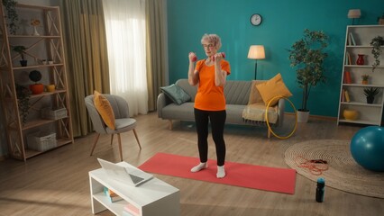 Elderly woman exercising with dumbbells in front of a laptop in the living room. A woman watches a video lesson on a laptop and repeats the exercise on the muscles of the arms, biceps.