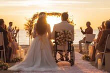 Back view of a couple of disabled man in wheelchair and elegant woman in weeding dress celebrating celebrating their wedding on the beach at sunset with friends and family.