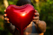 Close de coração vermelho metalizado sendo oferecido, segurado pelas mãos de uma mulher jovem na frente de seu rosto com fundo de vegetação desfocado.