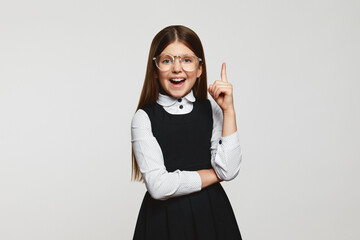 Wall Mural - Surprised smart little schoolgirl wearing eyeglasses and school uniform pointing up with open mouth while having idea against white background