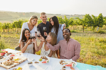 Happy adult friends having fun drinking red wine and eating together with vineyard in background - Multiracial people doing party at summer time in countryside resort - Main focus on faces