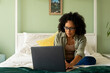 Biracial woman doing paperwork using laptop and smartphone on bed in bedroom