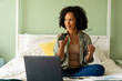 Biracial woman doing paperwork using laptop and talking on smartphone on bed in bedroom