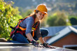 Smiling female technician in work clothes, helmet and with a tool installs solar panels on the roof of a modern house on a sunny day. The concept of modern technology using renewable energy