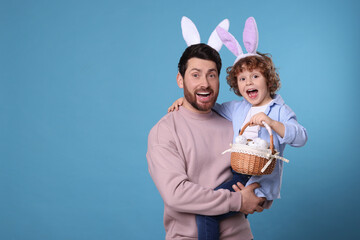 Canvas Print - Happy father and son in cute bunny ears headbands on light blue background. Boy holding Easter basket with painted eggs, space for text