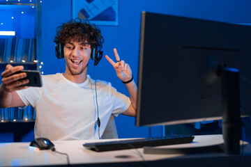 Arabic guy gamer in a headset playing computer games sits at a computer at night and makes a video call on a smartphone, shows a greeting gesture