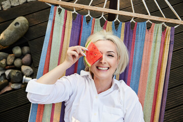 Happy adult woman having fun lying on hammock in backyard terrace eating watermelon