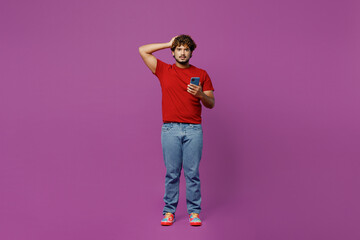Wall Mural - Full body young shocked frightened unhappy Indian man in red t-shirt casual clothes hold head use mobile cell phone look camera isolated on plain purple background studio portrait. Lifestyle concept