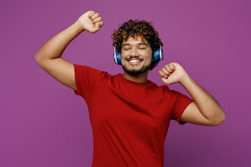 Wall Mural - Young cheerful smiling happy Indian man he wears red t-shirt casual clothes listen to music in headphones dance raise uo hands isolated on plain purple background studio portrait. Lifestyle concept.