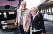 © luciano - Smiling senior white haired couple of passengers with trolley and suitcases out the airport. Old senior man and woman leaving for vacation. Arrival or departure